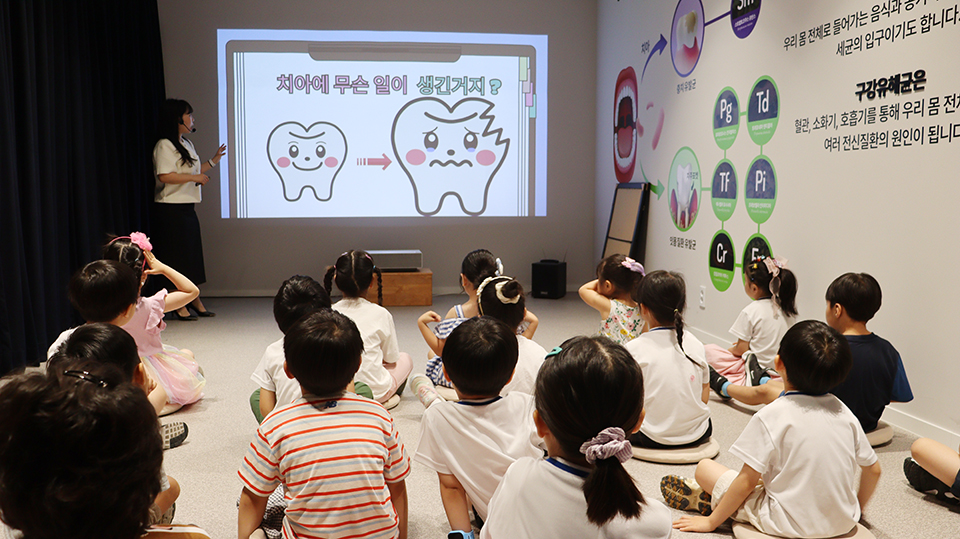 유치원 아이들이 사과나무치과병원 1층 영상관에서 치아 건강의 중요성에 관해 설명을 듣고있다.