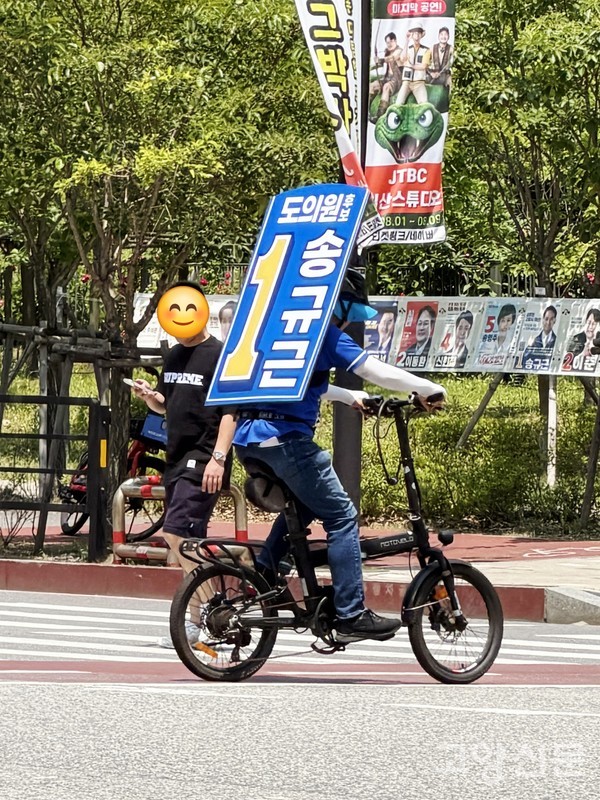 등 뒤에 큼지막한 '워킹보드'를 메고 자전거로 지역구 횡단보도를 건너며 지지를 호소하고 있다.