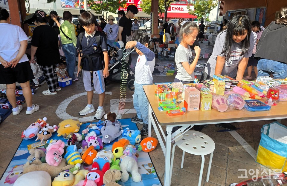 바자회 장난감 코너에 모인 아이들이 알록달록한 인형과 장난감들을 호기심 어린 눈빛으로 살펴보며 즐거운 시간을 보내고 있다.